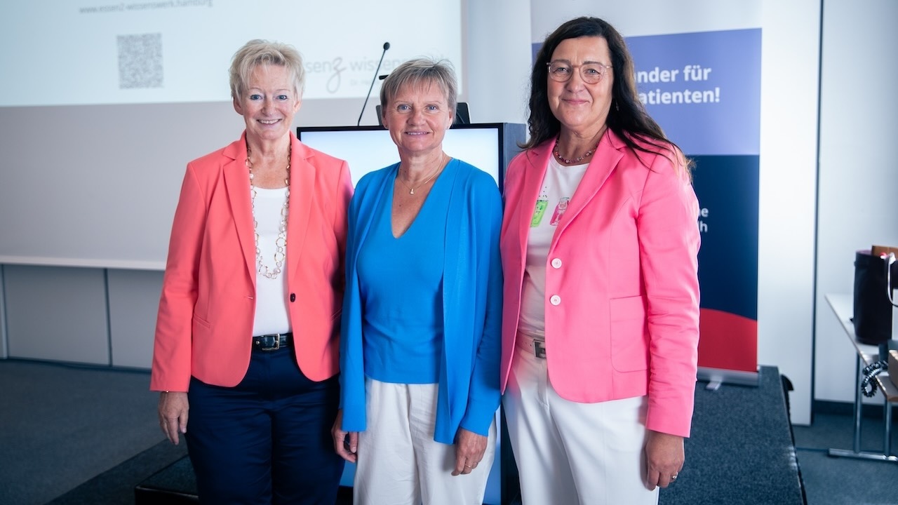 von links stehen Sylvia Fresmann in lachsfarabenem Blaazer, Mirija Krauß in blauer Strickjacke und Heike Wilken in rosafarbenem Blazernebeneinander neben der Bühne der DGDH Jahrestagung 2025