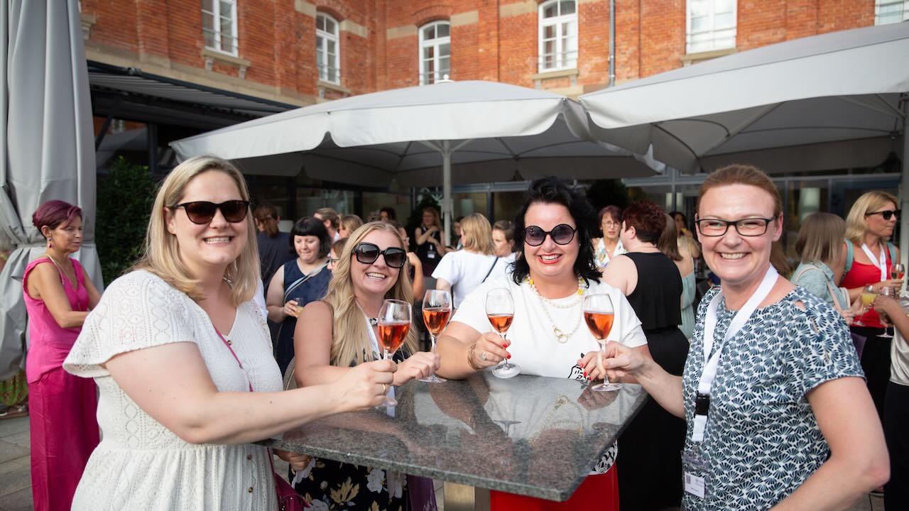 Vier Frauen stehen an einem Stehtisch und prosten mit orangem Getränk lächelnd in die Kamera, im Hintergrund zu sehen sind die anderen Teilnehmerinnen und Teilnehmen der DGDH Jahrestagung 2025 überwiegend unter großen Sonnenschirmen