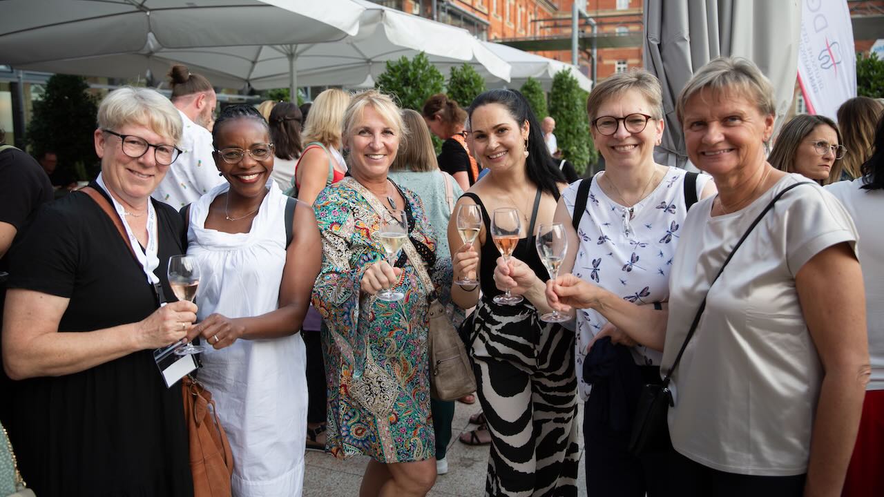 Sechs Frauen stehen im Halbkreis und prosten mit orangem Getränk lächelnd in die Kamera, im Hintergrund zu sehen sind die anderen Teilnehmerinnen und Teilnehmen der DGDH Jahrestagung 2025, überwiegend unter großen Sonnenschirmen