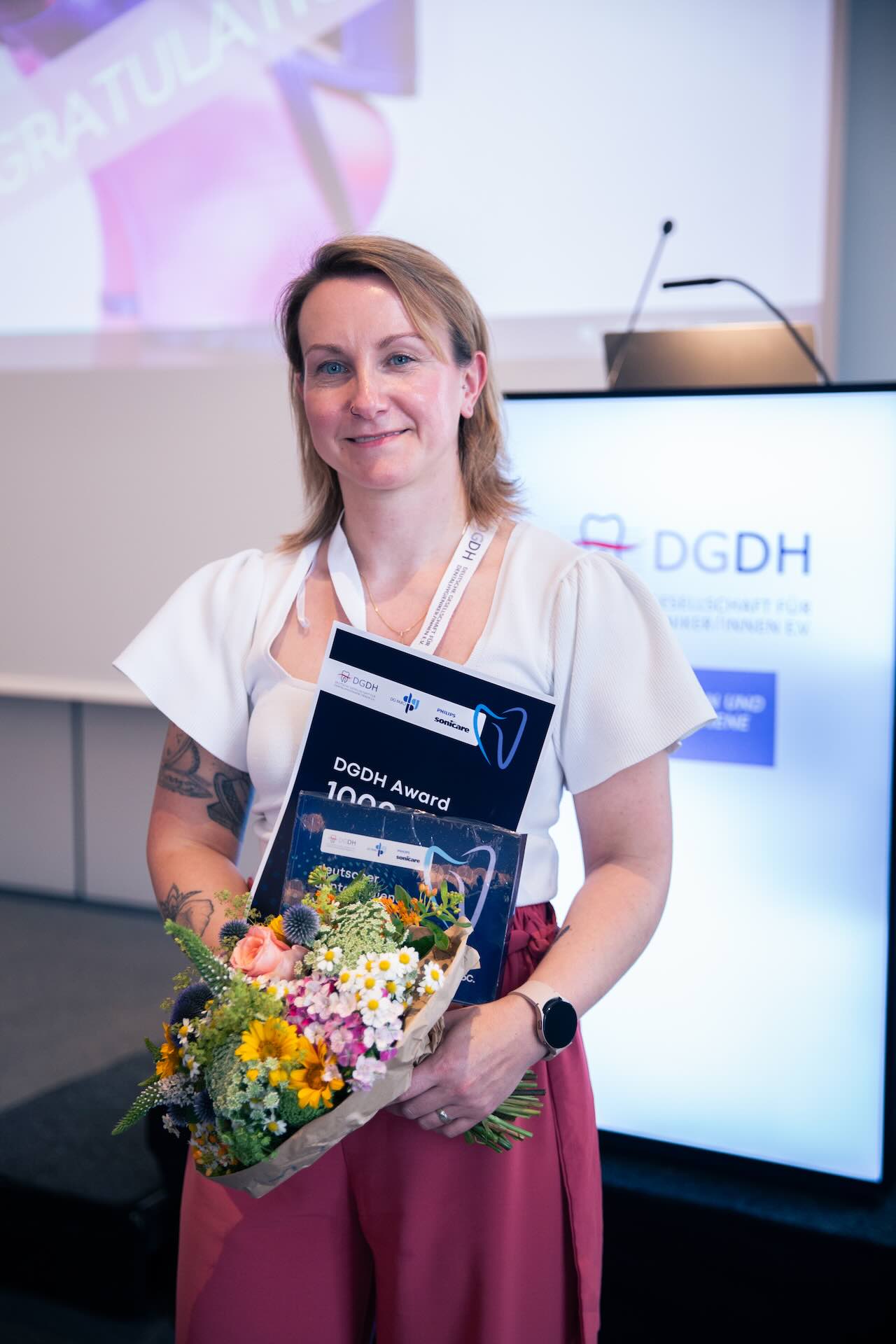 Magdalena Frackiewicz steht mit ihrer Urkunde und einem bunten Blumentstrauß neben der Bühne der DGDH Jahrestagung 2025