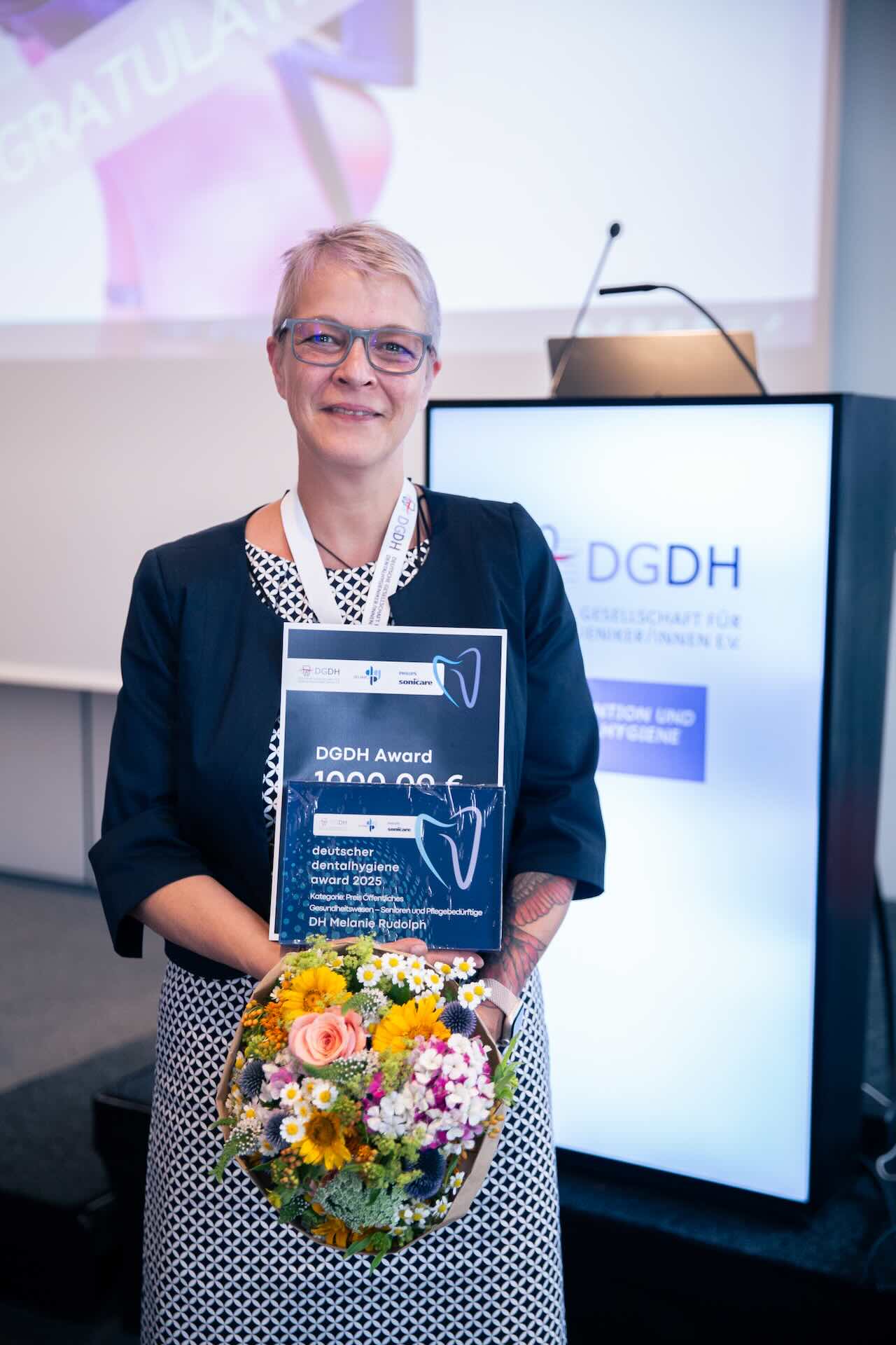 Melanie Rudolph steht mit ihrer Urkunde und einem bunten Blumentstrauß neben der Bühne der DGDH Jahrestagung 2025