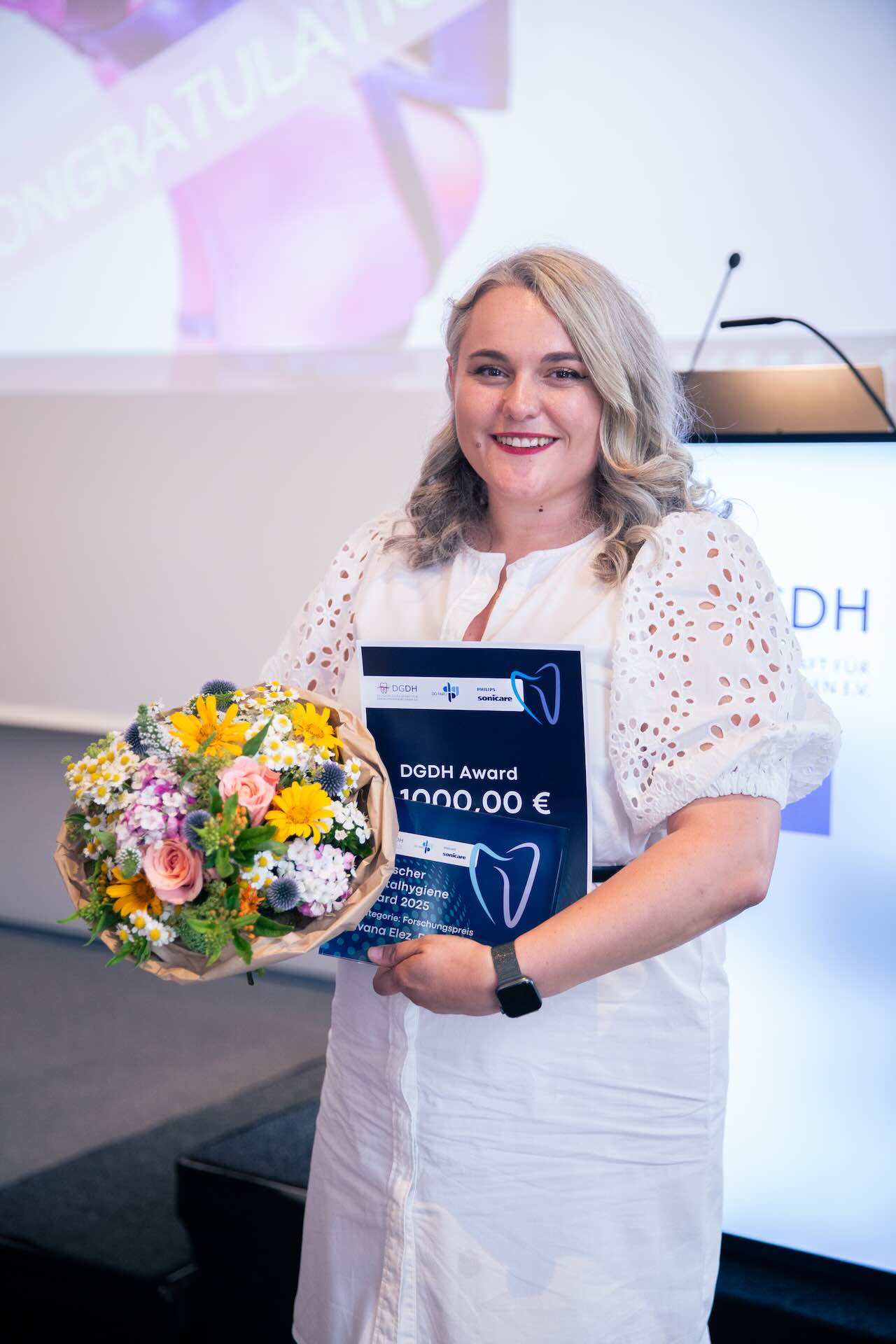 Ivana Elez steht mit ihrer Urkunde und einem bunten Blumentstrauß neben der Bühne der DGDH Jahrestagung 2025