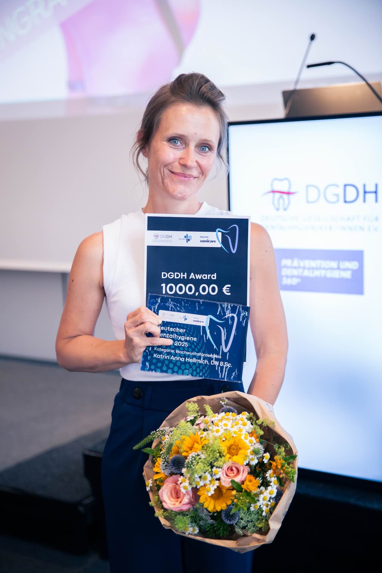Katrin Hellmich steht mit ihrer Urkunde und einem bunten Blumentstrauß neben der Bühne der DGDH Jahrestagung 2025