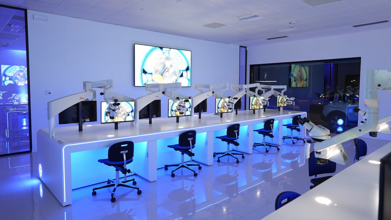 Übungsraum der Komet Academy in Verona mit zehn Arbeitsplätze, ausgestattet mit Leica M320 4K-Mikroskopen und vollständig eingerichteten Dental-Simulationseinheiten. Der Raum ist indirekt mit blauem Licht ausgeleuchtet, im Boden spiegeln sich die Tische und Stühle wider. An einer Wand ist ein großer Monitor befestigt, die Wand daneben hat ein großes schwarzes Fenster, in dem sich die Einrichtung widerspiegelt.