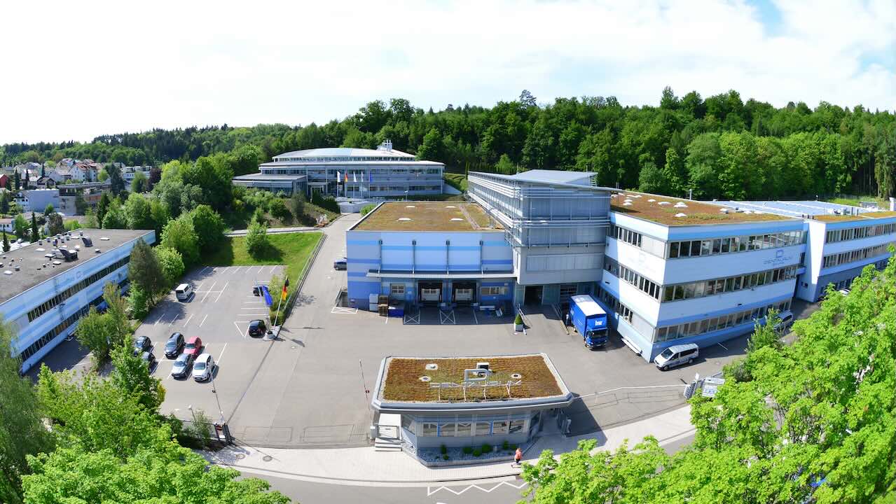 Die Dentaurum Firmenzentrale beginnt mit einem Pförtnerhäuschen, links und rechts davon sind Schranken. Dahinter sind sechs weitere Gebäude zu sehen.