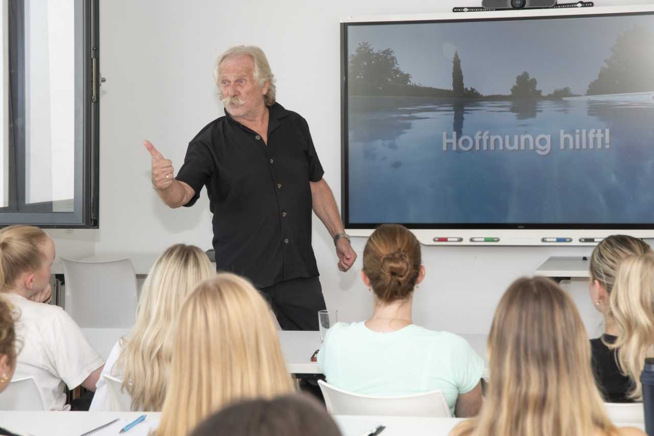 Ein Mann mit schwarzem Hemd steht vor einem Bildschirm mit einem Flussbild. Im Vordergrund Zuhörerinnen und Zuhörer.
