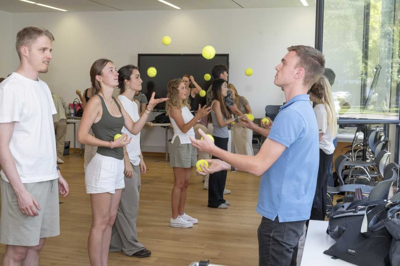 Junge Menschen in Sportkleidung stehen in einer Sporthalle und jonglieren mit gelben Tennisbällen