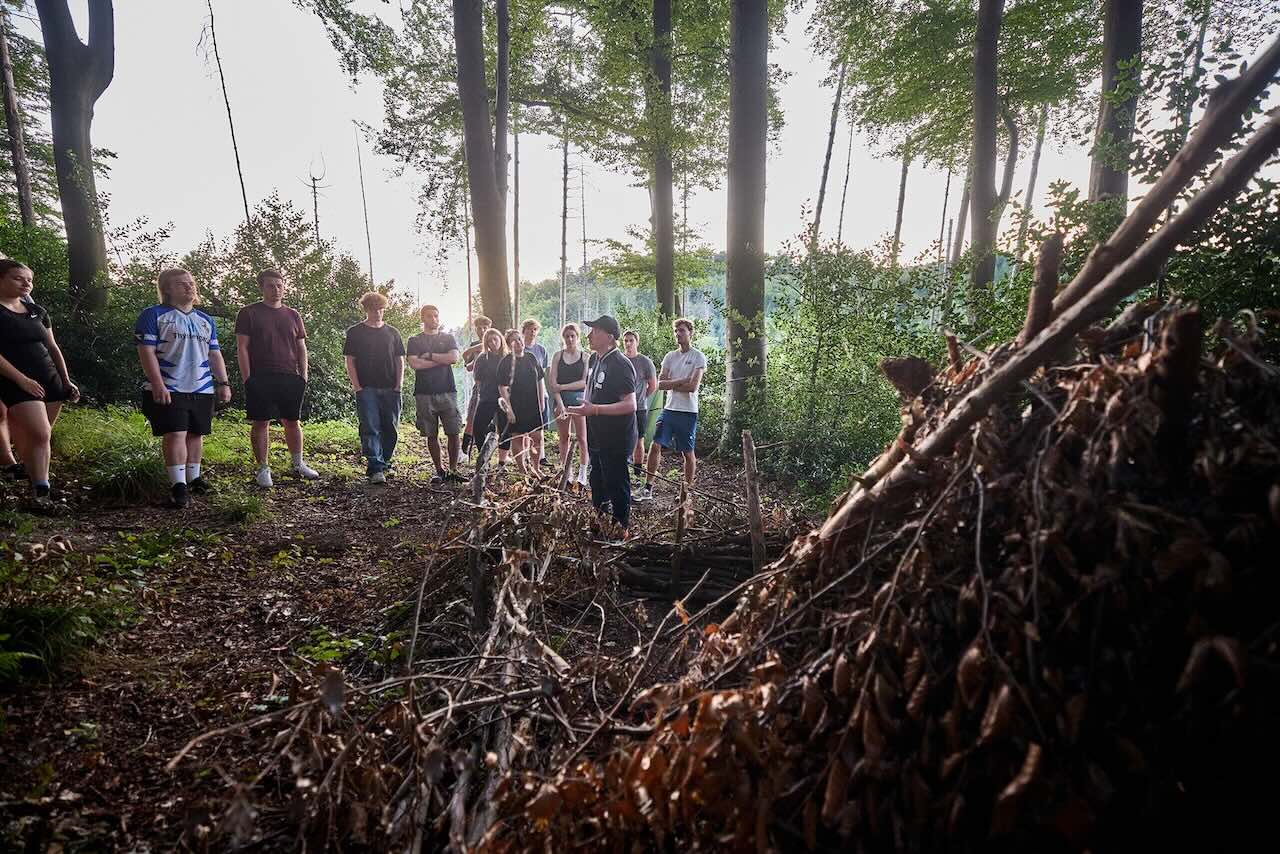 Mehrere Menschen stehen in einem lichten Wald, rechts im Bild aufgeschichtete Äste.
