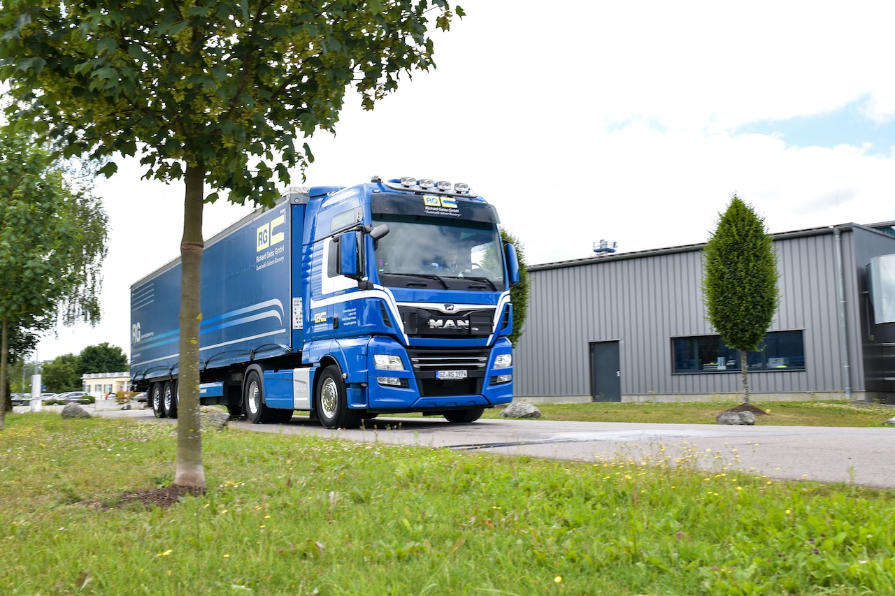 Ein blauer LKW vor einem weißen Flachdachgebäude, davor Wiese und ein kleiner Baum.