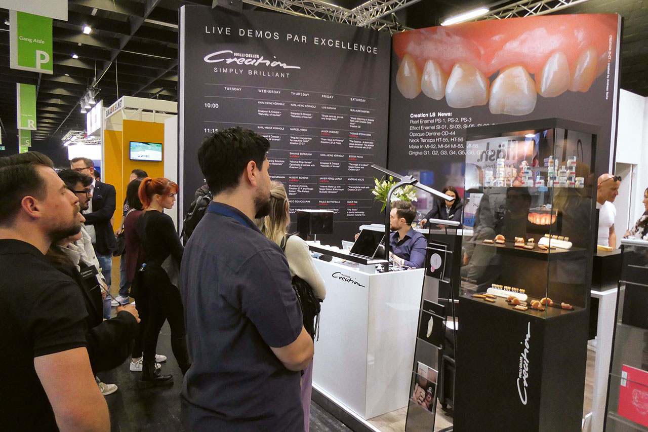 Menschen im Vordergrund vor einem Messestand schauen bei einer Live-Demonstration zu, rechts im Bild eine schwarze Vitrine mit verschiedenen Zahnersatzbeispielen.