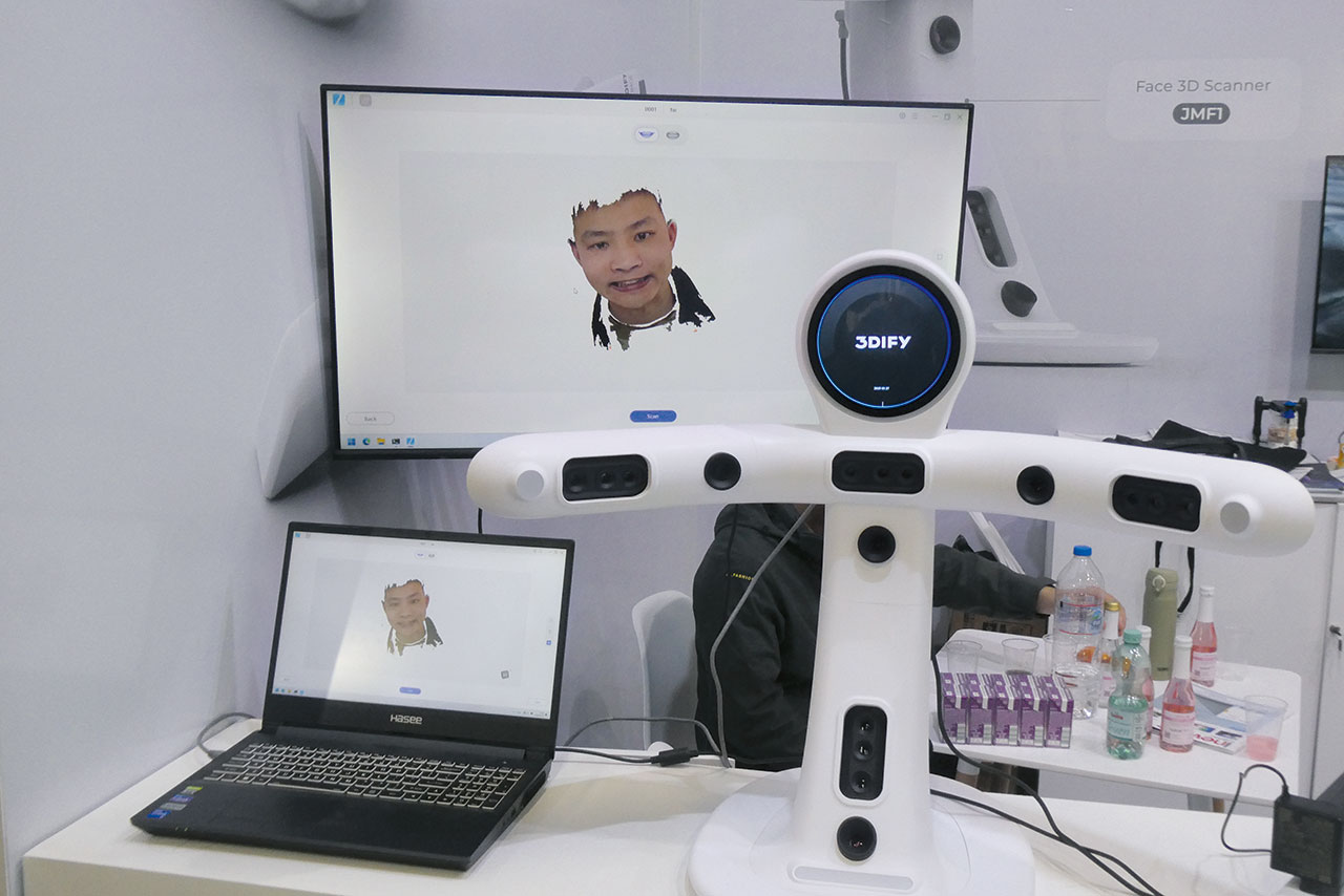 Auf einem Tisch steht links ein kleines Notebook, darüber ein Bildschirm, der ein gescanntes Gesicht zeigt, daneben ein Gesichtsscanner, im Hintergrund ein Arbeitsplatz mit Getränken.