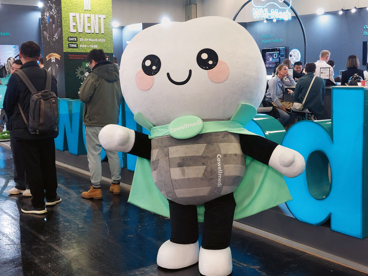 Ein als Maskottchen verkleideter Mensch vor einem Messestand mit großem niedlichem Gesicht und grünem Umhang