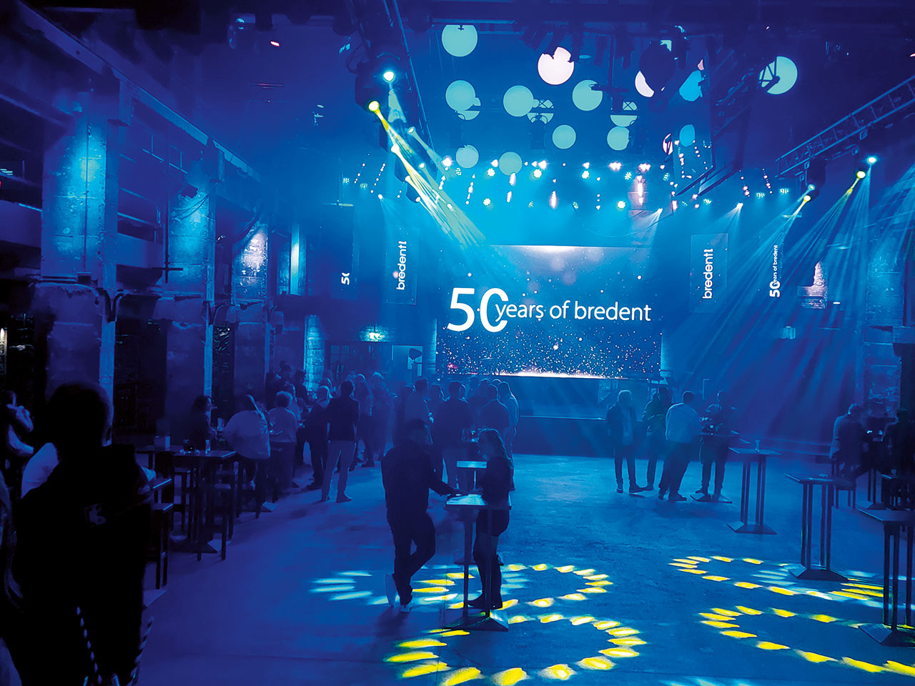 Ein in blaues Licht getauchter Veranstaltungsraum im Hintergrund ein Schild auf dem steht „50 years of bredent“, Menschen an Stehtischen.