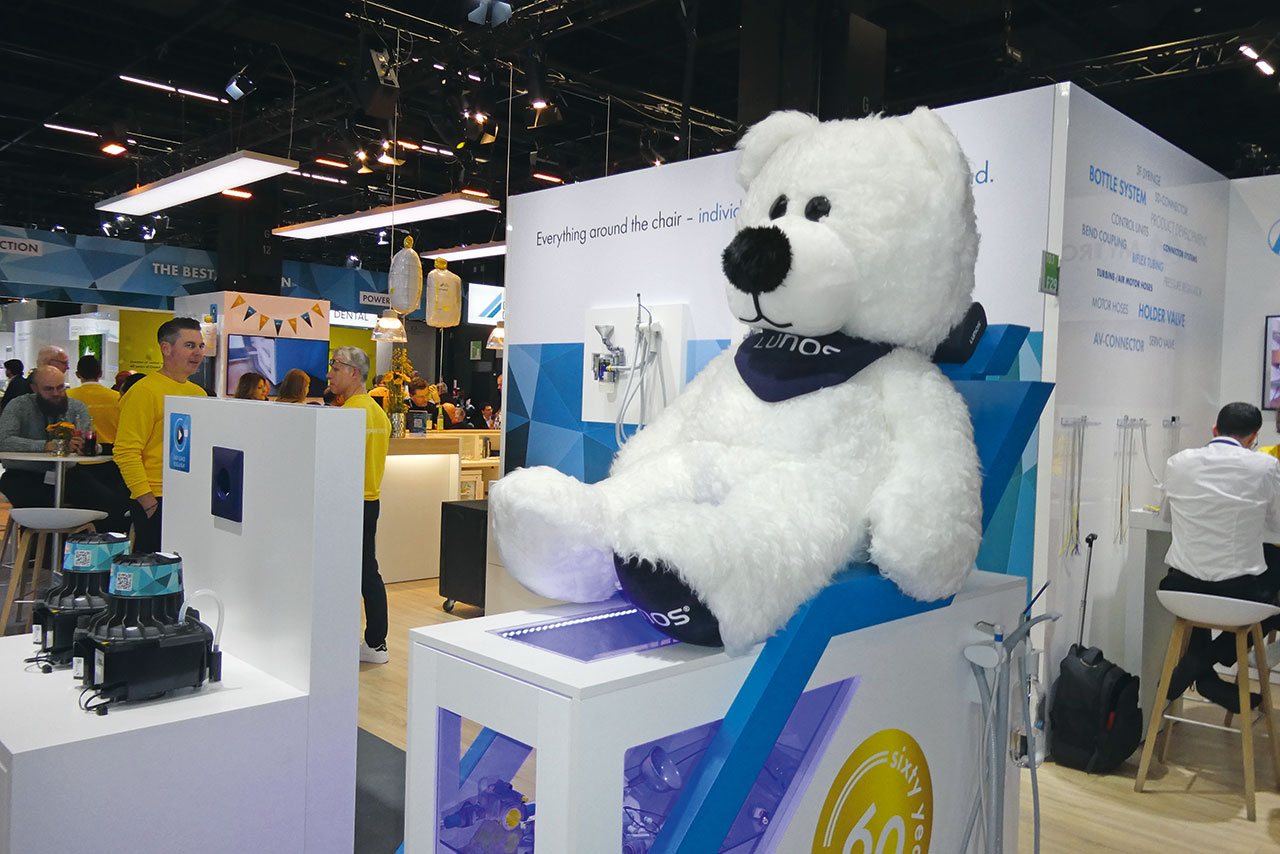 Ein großer weißer Plüschbär mit dunklem Halstuch auf dem „Lunos“ steht, lehnt gegen ein Regal am Stand von Dürr Dental