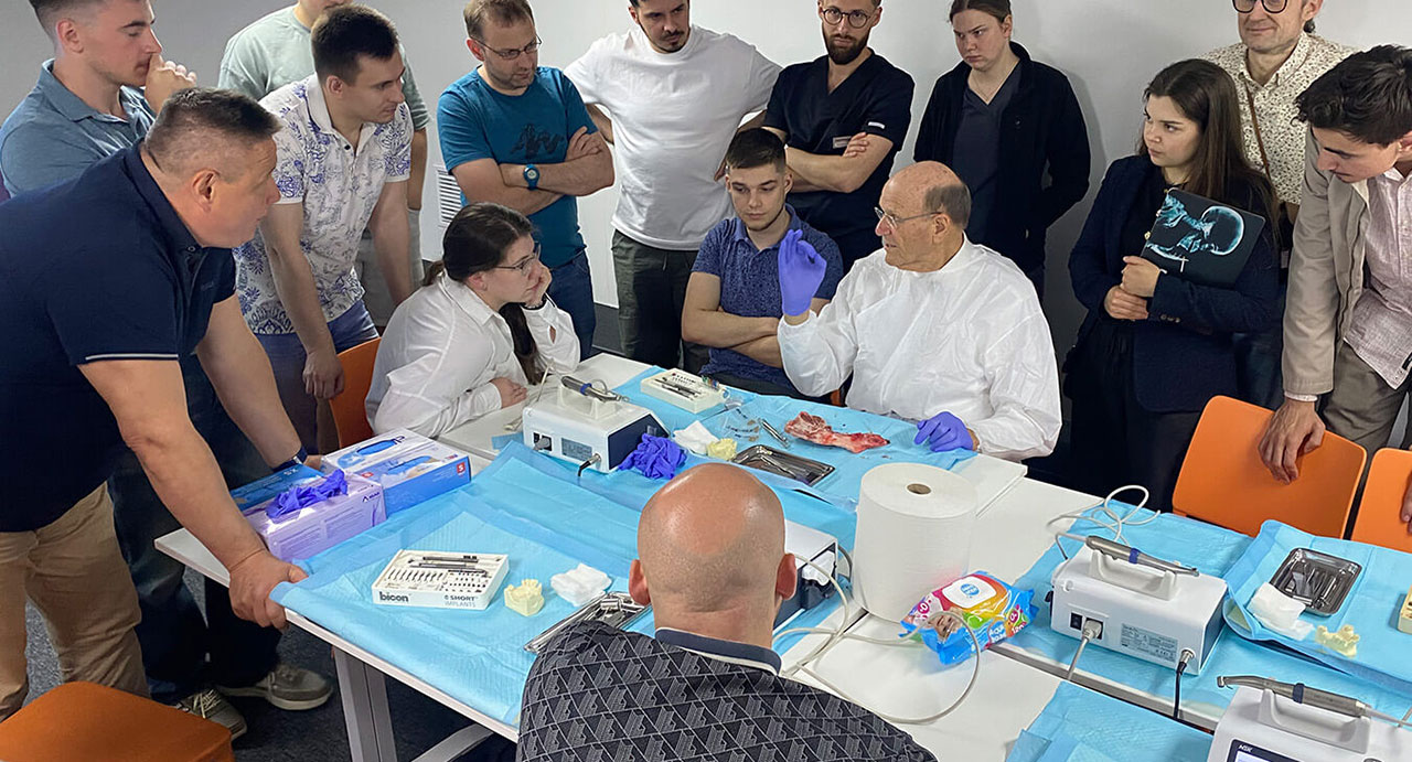 Um einen rechteckigen blauen Tisch , auf dem chirurgische Instrumente und Materialien liegen, stehen ca. zehn Männer und Frauen. Prof. Ewers sitzt am Tisch und zeigt etwas an einem Stück Schweinekiefer, das vor ihm liegt.