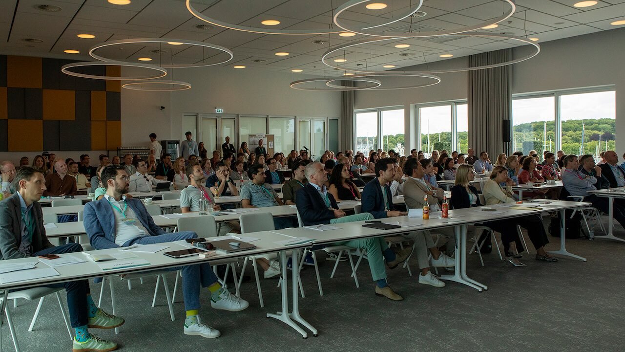 Blick in einen gut gefüllten Vortragsraum, in dem Menschen an Tischen sitzen und einem Vortrag folgen.