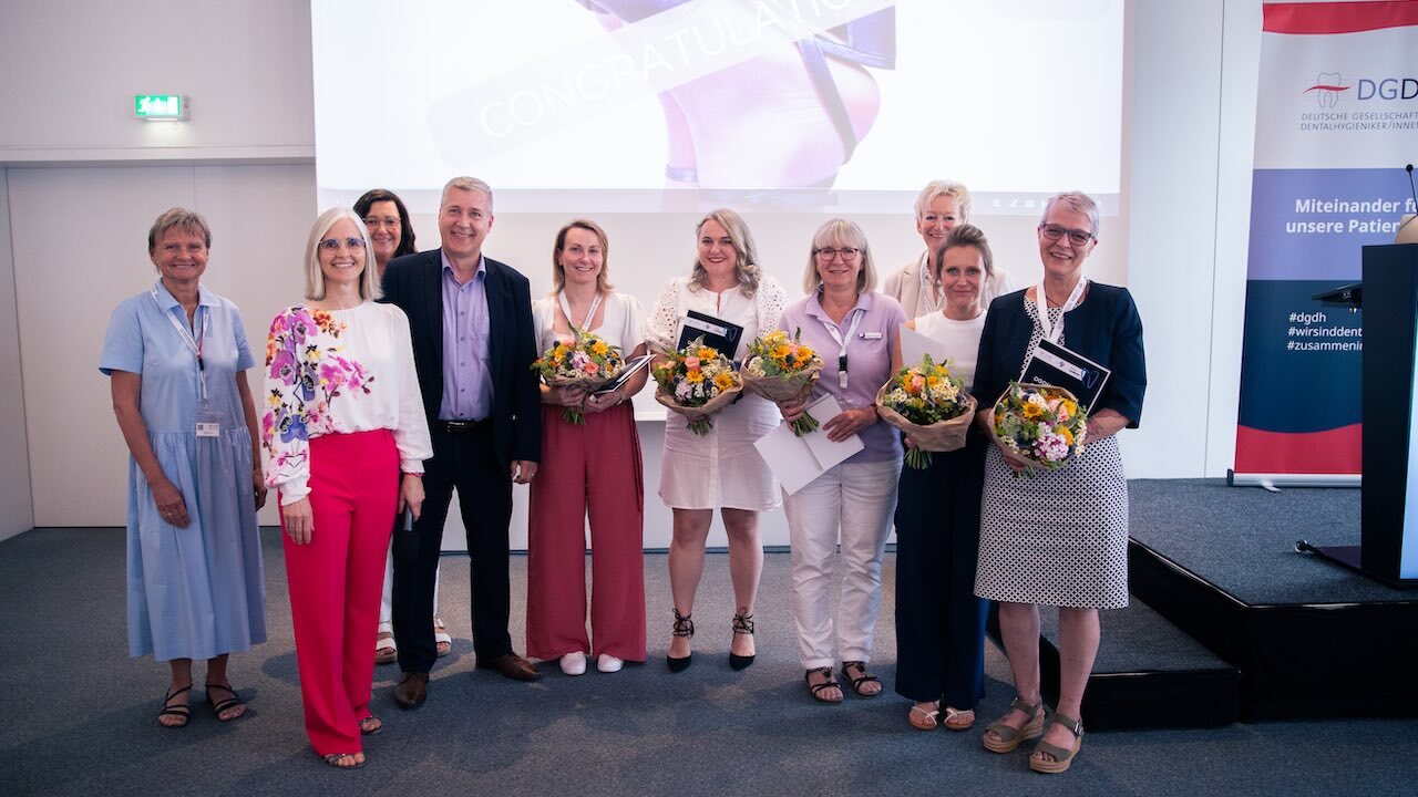 Die Sponsoren des Deutschen Dentalhygiene Awards und die fünf Gewinnerinnen der Awards neben der Bühne der DGDH-Jahrestagung. Die fünf Gewinnerinnen halten alle ihre Urkunde und einen bunten Blumenstrauß in den Händen.
