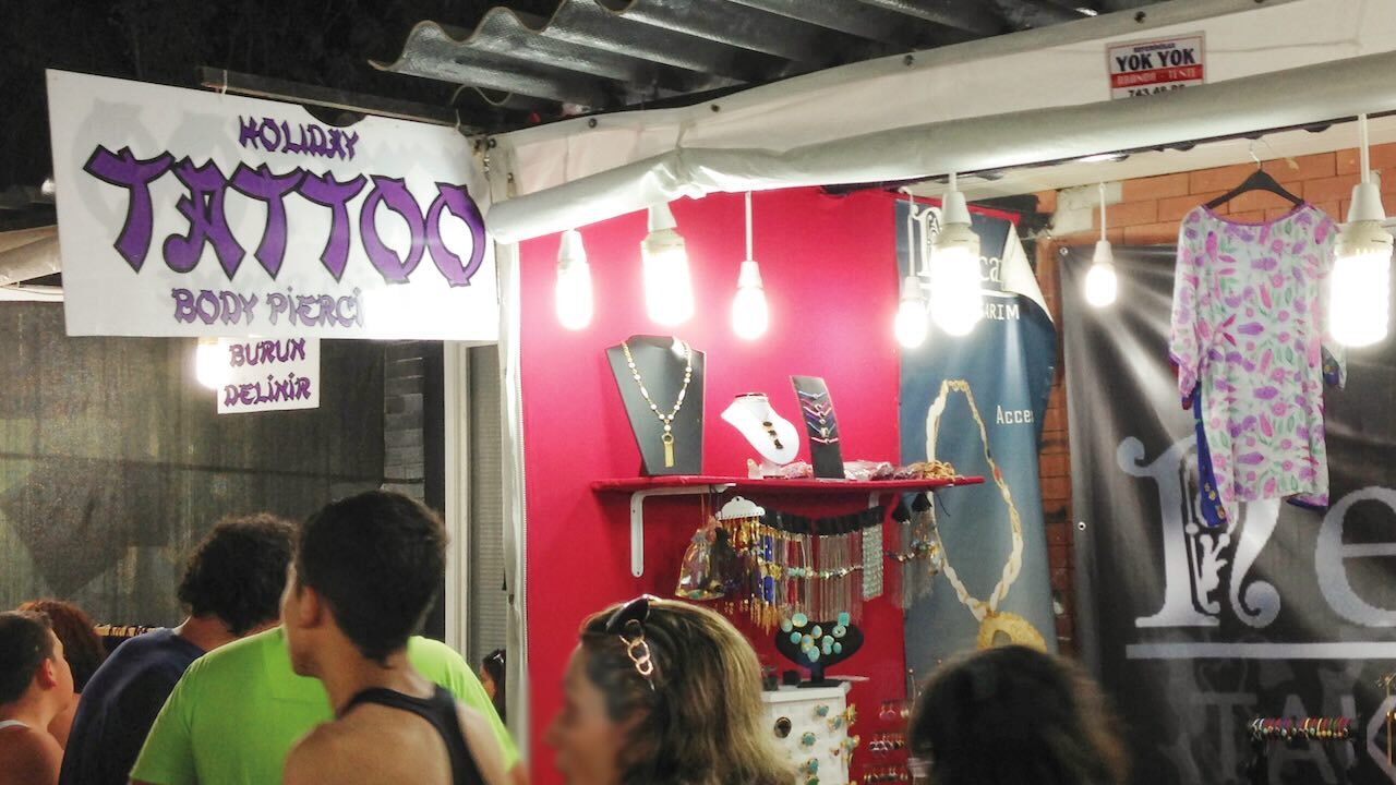 Werbung für ein Tattoo-Studio an einem Verkaufsstand mit Wellblechdach in einer belebten Straße. Es ist Abend und an dem Verkaufsstand sind Schmuck und Textilien zu sehen, es leuchten mehrere Glühbirnen an dem Verkaufsstand, da die Umgebung dunkel ist.