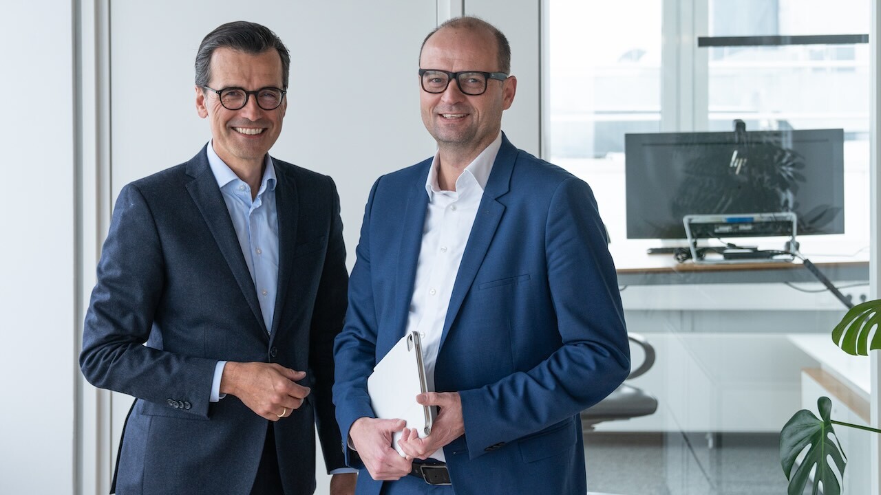 Links steht Stephan Köhler, Sprecher der Geschäftsführung von Brasseler. Er trägt eine Brille, einen dunkelblauen Anzug und ein hellblaues Hemd. Rechts neben ihm steht Carsten Cieslik, Leiter der Business Unit Komet Dental. Er trägt Brille, einen mittelblauen Anzug und ein weißes Hemd. In der Hand hält er ein silbernes Tablet. Im Hintergrund ist ein kleiner Blick in ein Büro mit Monitor, Schreibtisch und Bürostuhl.