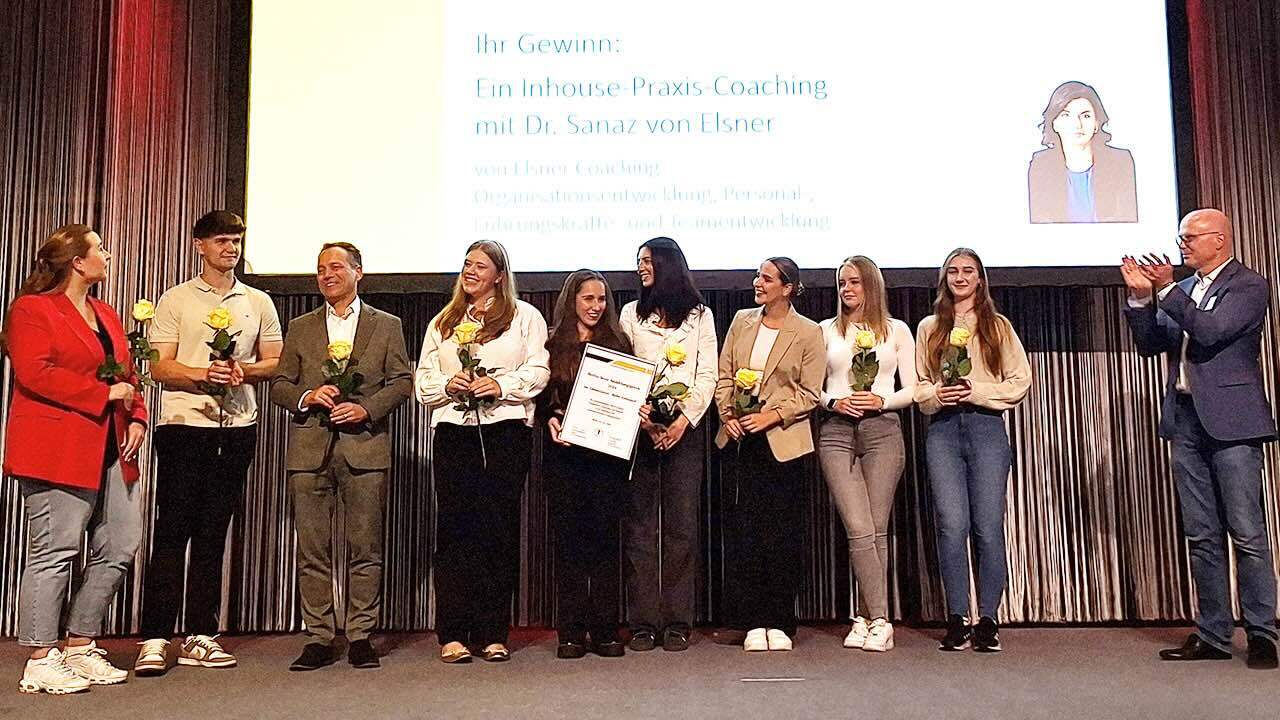 Der Gewinner Dr. Robert Heym steht mit einem männlichen und sieben weiblichen Teammitgliedern in einer Reihe, alle haben eine gelbe Rose in der Hand, eine junge Frau hält die eingerahmte Urkunde. Links davon steht Dr. Karsten Heegewaldt, Präsident der ZÄK Berlin, und applaudiert.