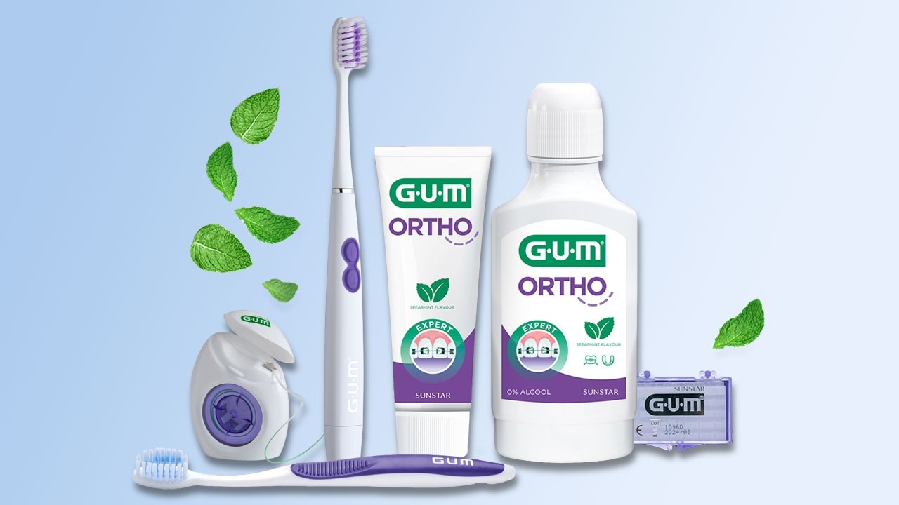 Nebeneinander stehen Gum Ortho Floss, die Gum Sonic Ortho Schallzahnbürste, das Gum Ortho Zahngel und die Gum Ortho Mundspülung, das Gum Ortho Wachs; davor liegt die Gum Ortho Zahnbürste. Über dem Floss sind fünf Minzblätter und über dem Wachs ein Minzblatt zu sehen.