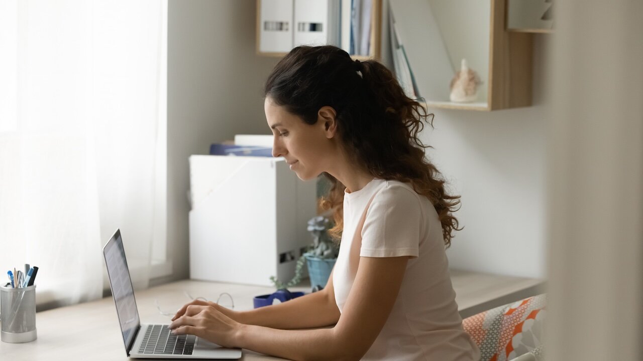 Eine junge Frau sitzt zu Hause an ihrem Schreibtisch und tippt in ihr Laptop. An der Wand rechts neben ihr hängen Regale mit Unterlagen, auf dem Schreibtisch sind Unterlagen, ein Blumentopf mit verschiedenen Blumen und ein Metallgefäß mit Stiften. Neben ihren Händen liegt eine Brille. Sie trägt ein weißes T-Shirt und hat lange dunkle Haare, die sie nach hinten gebunden hat.