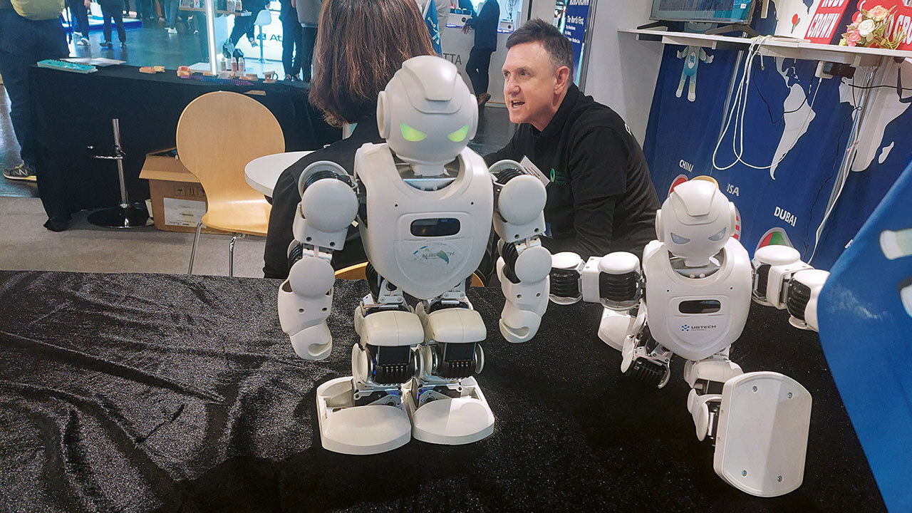 Zwei Roboter im Vordergrund mit menschenähnlicher Figur stehen beziehungsweise sitzen auf einem Tisch an einem Messestand, dahinter sitzt ein Mann.