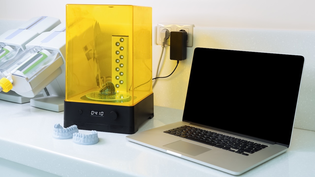 Zwei zahntechnische Modelle auf einem Labortisch vor einem 3D-Drucker, rechts daneben ein aufgeklapptes Notebook mit dunklem Bildschirm.