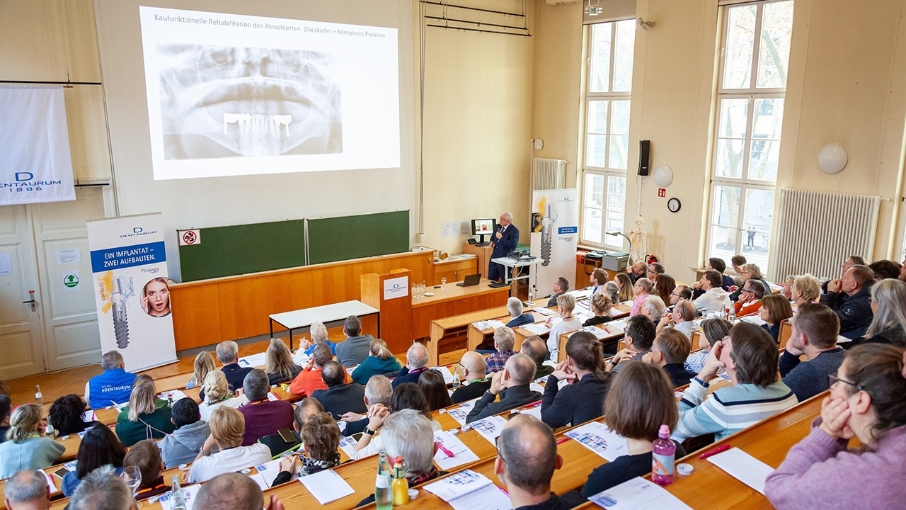 Foto zeigt Menschen in einem Hörsaal sitzend, die dem Referenten vorne rechts zuhören, über der grünen Tafel ist eine Leinwand mit einem Röntgenbild eines Ober- und Unterkiefers zu sehen. Links und rechts von der Tafel stehen Aufsteller der Firma Dentaurum.