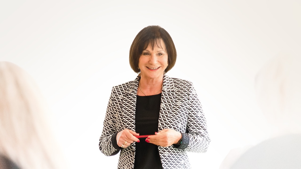 Sylvia Wuttig schaut lächelnd an der Kamera vorbei. Sie trägt ein schwarzes T-Shirt unter einem schwarz-weißen Blazer und hält etwas rotes Schmales in der Hand, das man nicht erkennen kann. Der Hintergrund ist weiß. In der linken unteren Ecke lassen sich verschwommen lange blonde Haare erkennen.