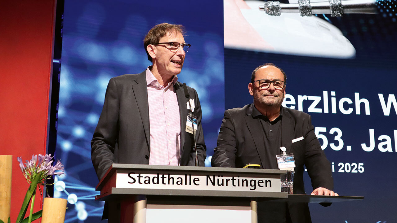 Zwei Männer stehen hinter einem Rednerpult der Stadthalle Nürtingen auf der Bühne mit einem roten Vorhang (links).