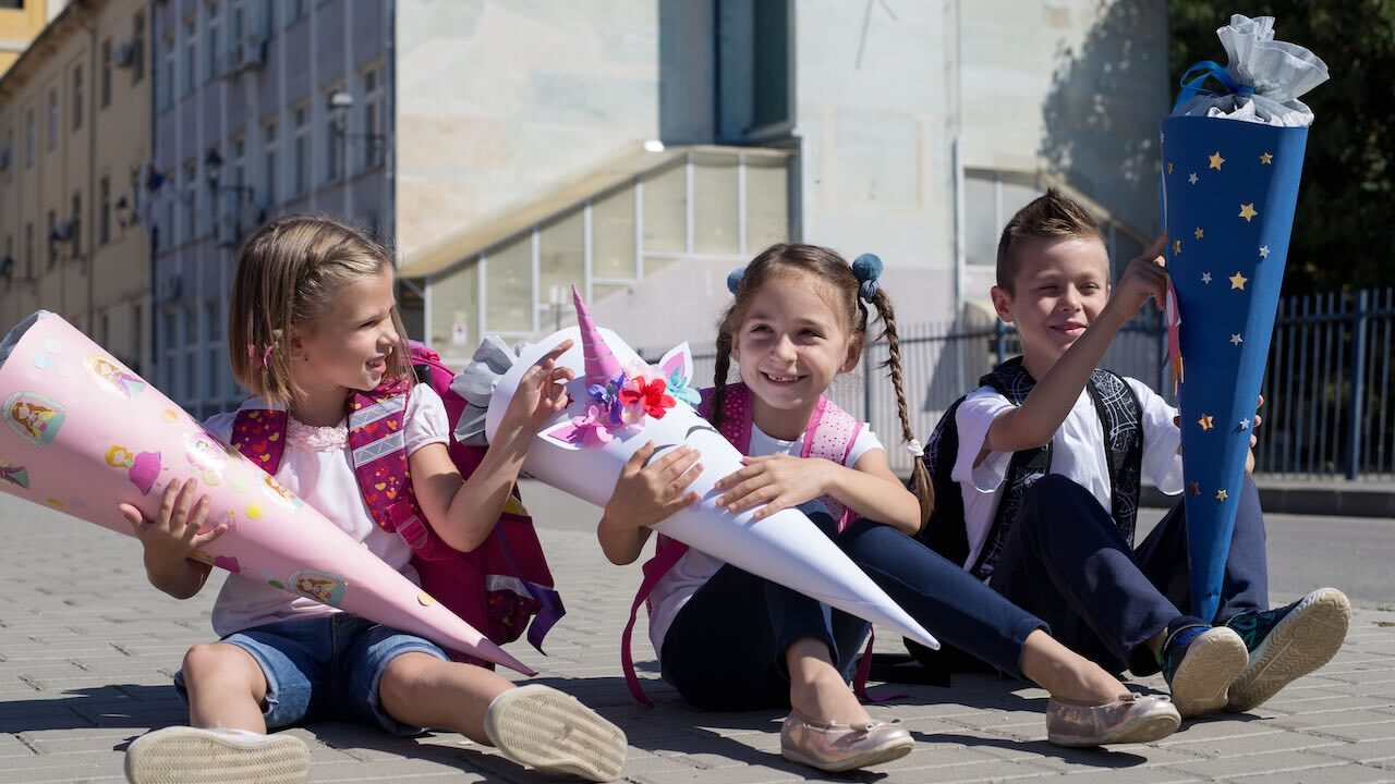 Von links nach rechts sitzen zwei Mädchen und ein Junge draußen auf dem Boden. Im Hintergrund sieht man zwei Gebäude. Das linke Mädchen hält eine rosafarbene Schultüte mit Prinzessin-Lillifee-Aufklebern, das Mädchen in der Mitte eine weiße Schultüte mit einem aufgeklebten fantasievollen Elefantengesicht und der Junge eine dunkelblaue Schultüte. Alle drei sind sommerlich angezogen mit Kurzarm-T-Shirts.