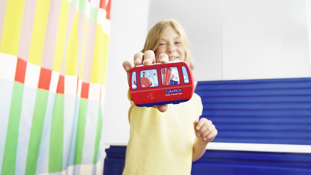 Ein Mädchen mit blonden langen Haaren im hellgelben T-Shirt hält vor sich ein kids Travel Set mit einer roten Bandrole, Motiv Gondel, in die Kamera. links neben ihr ist ein langer bunter Vorhang zu sehen, hinter ihr eine weiß-blaue Wand.