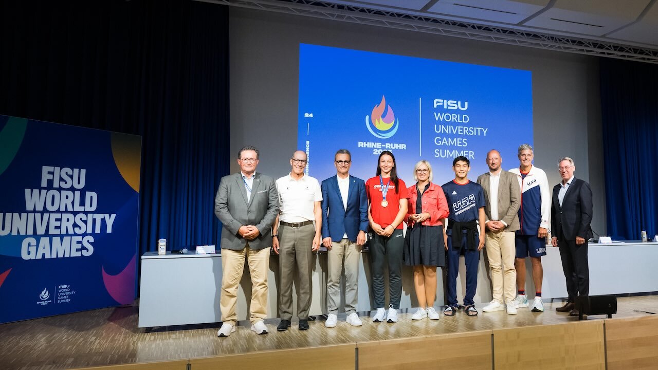 In einer Reihe stehen neun Personen auf der Bühne der Abschlusspressekonferenz der FISU World University Games Rhine-Ruhr 2025, deren Logo im Hintergrund auf einer großen Leinwand zu sehen ist. An vierter Stelle von link steht Anna Monta Olek, die sich die Goldmedaille Judo in der Klasse bis 78 kg erkämpft hat. Sie trägt ein rotes Sportshirt, eine schwarze Trainingshose und weiße Sportschuhe, um den Hals hängt ihre Goldmedaille.