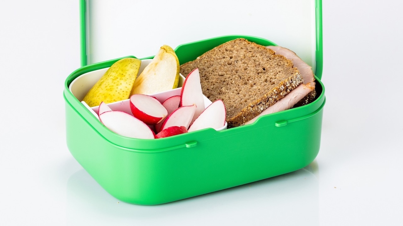 In einer offenen grünen Frühstücksdose liegen zwei Viertel Birne, einige in Spalten geschnittene Radieschen und zwei Scheiben Vollkornbrot, die mit einer Scheibe Wurst belegt ist. Die drei Inhalte sind durch weiße Platzteiler voneinander getrennt.