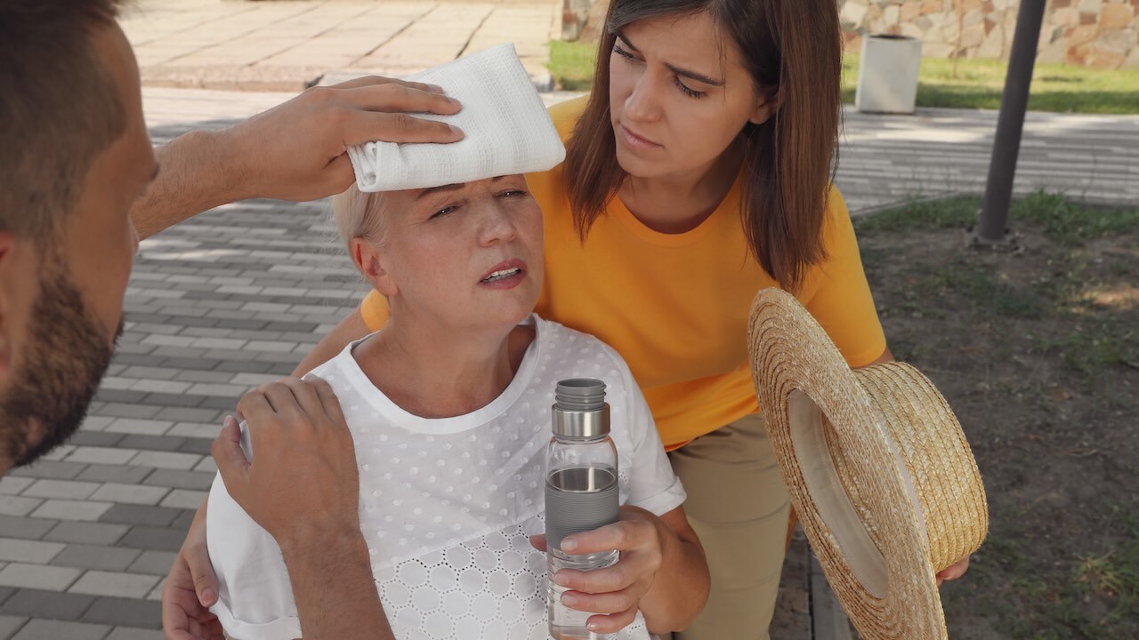 Eine blonde Frau sitzt erschöpft und hält eine Flasche mit Wasser in der linken Hand. Links beugt sich ein Mann zu ihr und hält ihr mit ein weißes Tuch an die Stirn. Rechts beugt sich eine junge Frau zu ihr und stützt ihr mit dem rechten Arm den Rücken. In der anderen Hand hält die junge Frau einen Strohhut.