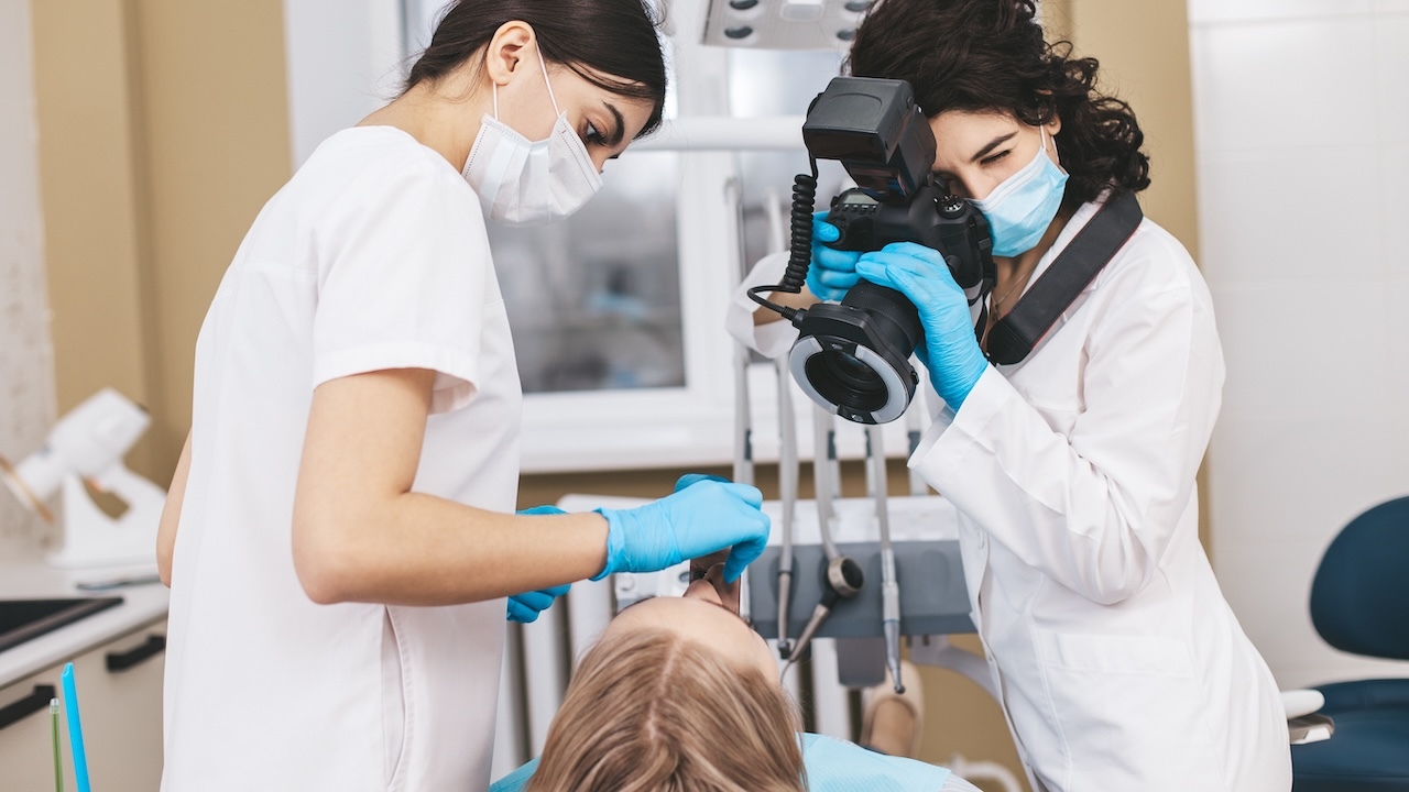 Zahnmedizinische Angestellte (links) und Zahnärztin mit Fotoapparat (rechts) stehen vor einer Behandlungseinheit, in der Mitte liegt eine Patientin auf dem Behandlungsstuhl.