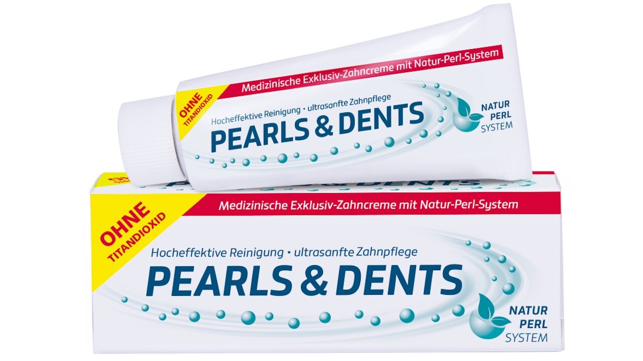 In der unteren Hälfte liegt längs ein Umkarton von Pearls & Dents, darauf liegt eine Tube der Zahnpasta. Auf der Verpackung und der Tube ist ein roter Vermerk „Ohne Titandioxid“ auf einem gelben Dreieck optisch hervorgehoben.