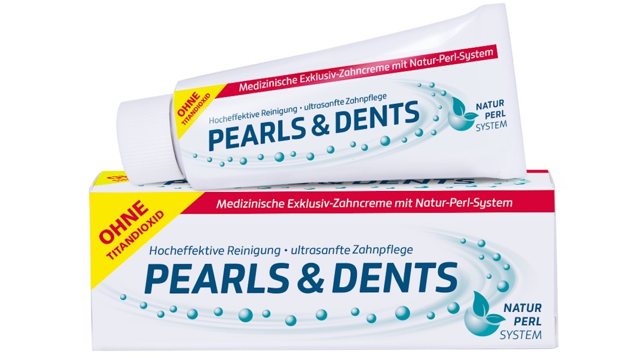 In der unteren Hälfte liegt längs ein Umkarton von Pearls & Dents, darauf liegt eine Tube der Zahnpasta. Auf der Verpackung und der Tube ist ein roter Vermerk „Ohne Titandioxid“ auf einem gelben Dreieck optisch hervorgehoben.