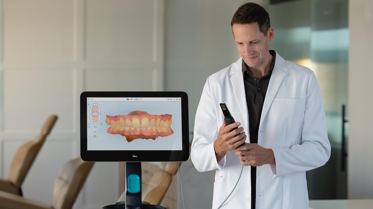 Ein Mann im weißen Kittel hält das Handstück des iTero Scanners in der Hand. Links neben ihm ist der Monitor zu sehen. Im Hintergrund sieht man drei Behandlungsstühle