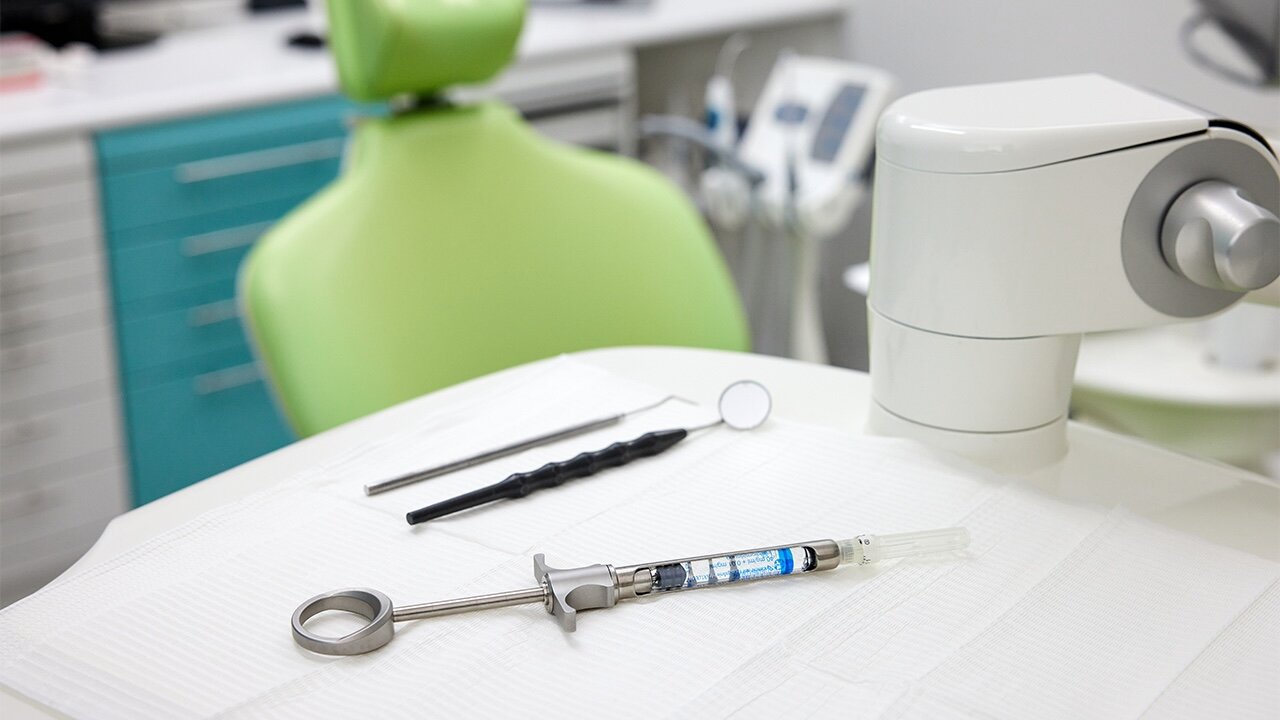 Auf einer zahnärztlichen Ablage liegen zwei Untersuchungsinstrumente und eine Spritze, im HIntergrund ein hellgrüner Patientenstuhl vor einem Schrank mit türkisen Schubladen.