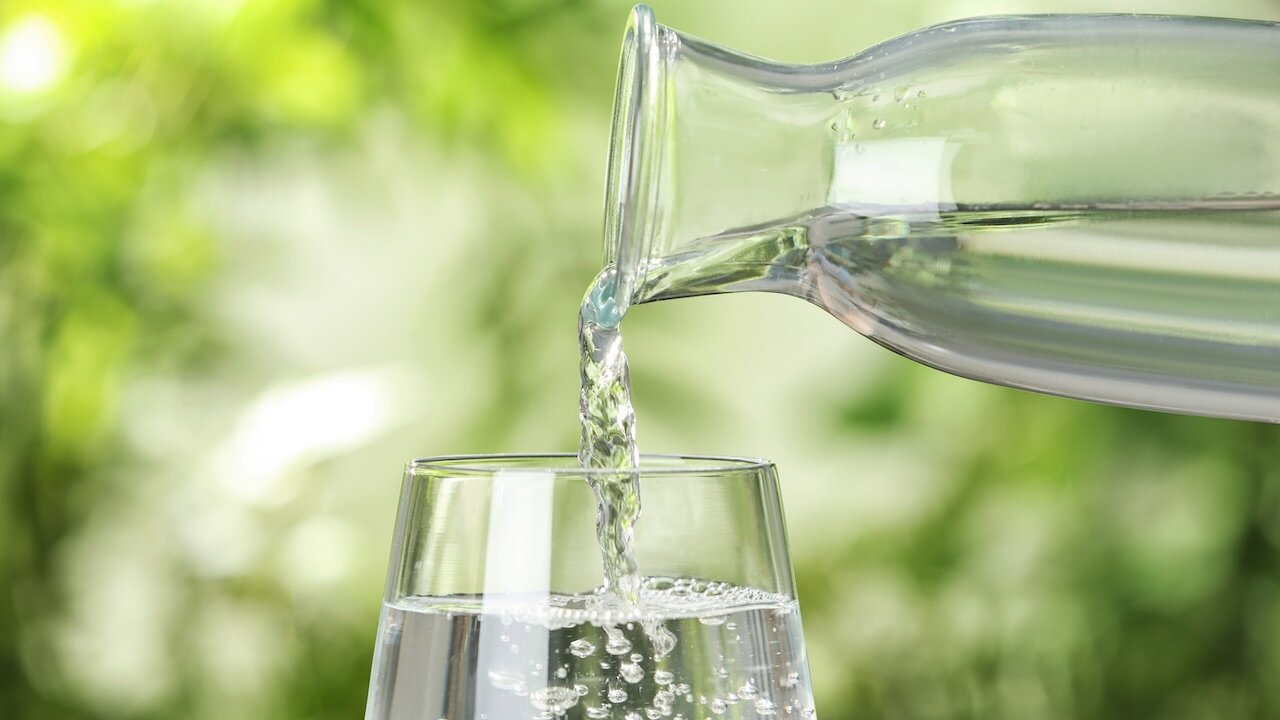 Aus einer Glaskaraffe fließt Wasser in ein Glas. Der Hintergrund zeigt verschiedene Grüntöne.