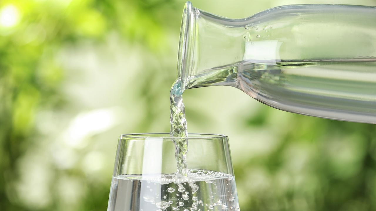Aus einer Glaskaraffe fließt Wasser in ein Glas. Der Hintergrund zeigt verschiedene Grüntöne.
