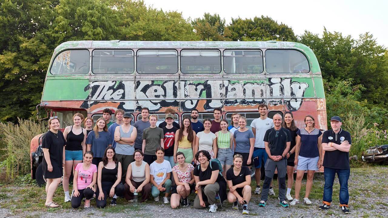 Eine Gruppe junger Menschen steht vor einem alten Bus mit der Aufschrift „Kelly Family“ vor Bäumen im Hintergrund auf einem Schotterweg.