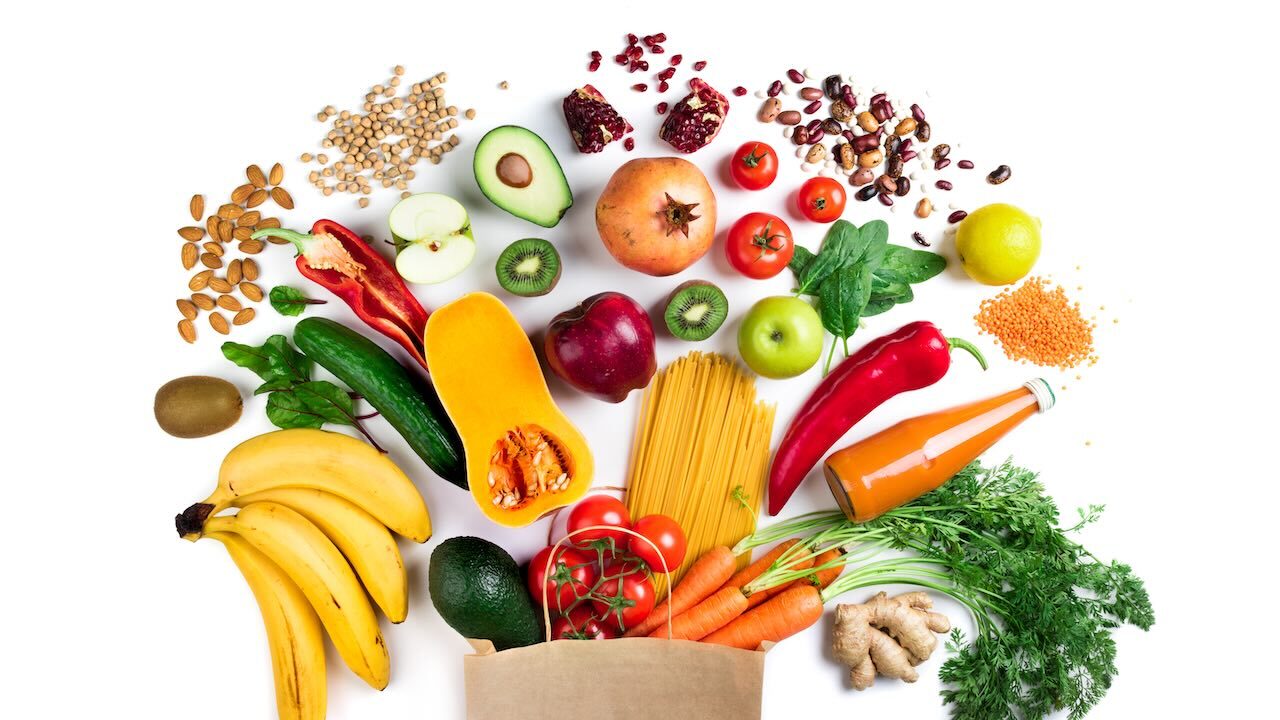 Aus einer Papiertüte schauen eine Avocado, Tomaten, Spaghetti und Möhren heraus. Drum herum liegen Bananen, rote Paprika, eine Zucchini, Äpfel, Kiwis, Mandeln, Kichererbsen, Granatapfelkerne und Granatapfelkerne, verschiedene Bohnensorten, und eine Flasche mit oranger Flüssigkeit.