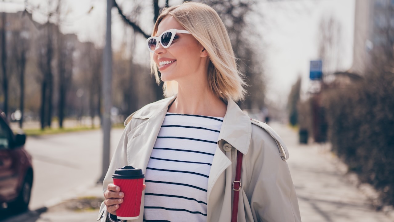 Eine junge Frau mit mittellangen blonden Haaren, einem leichten Pulli und einem hellen Trenchcoat steht lächelnd auf einem Bürgersteig. Sie trägt eine weiße Sonnenbrille und hält einen roten Trinkbecher in der Hand.