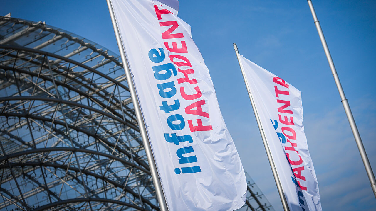 Zwei Flaggen mit blau und roter Aufschrift Infotage fachdental vor einem Drahtgeflecht und blauem Himmel