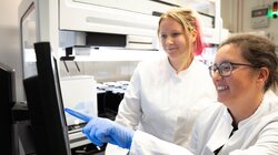 Die Wissenschaftlerin Ana Rita Brochado (rechts) und ihre Labormitarbeiterin Laura Sniegula blicken auf die Daten des Pipettierroboters. Beide lächeln und haben weiße Kittel an. Brochado richtet ihren Zeigefinger auf eine Stelle am Monitor.