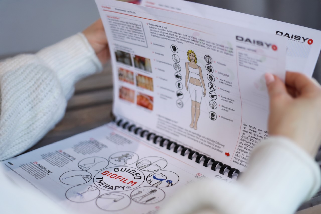 Frauenhände halten eine aufgeschlagene Webinarmappe in den Händen. Dort ist unter anderem eine Grafik zu sehen, die anzeigt, worauf sich Biofilm gesundheitlich auswirkt. Auf der gegenüberliegenden Seite ist die Grafik des GBT-Protokolls zu sehen. Die Schriften sind verschwommen.