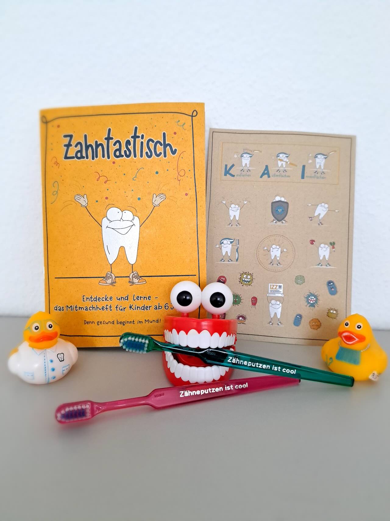 Das Heft Zahntastisch steht an eine helle Wand gelehnt neben einer schriftlichen Erklärung zur KAI-Methode. Davor sind links und rechts jeweils zwei Badeenten, die linke als Zahnarzt und die recht als ZFA. Dazwischen steht ein Klappergebiss, das eine grüne Handzahnbürste im zwischen den Zähnen hält, davor liegt eine rote Handzahnbürste.